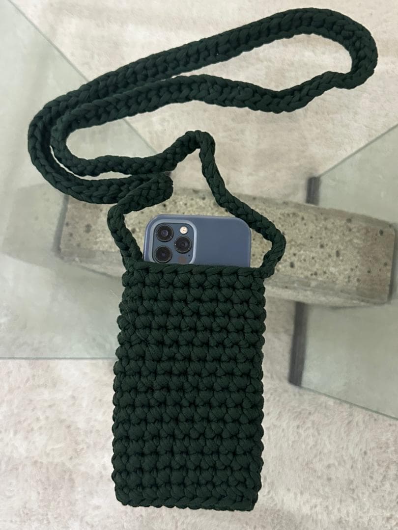 Pochette pour téléphone crochetée vert foncé à longue bandoulière, un téléphone glissé à l’intérieur.